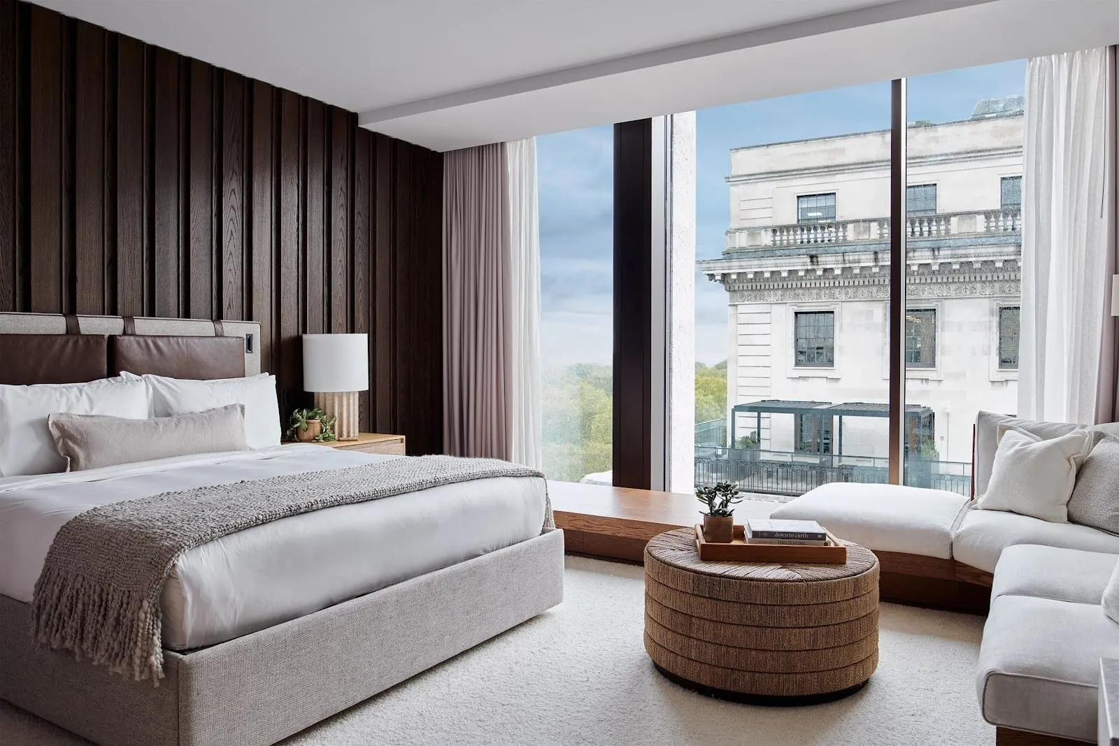 A 1 Hotel Mayfair bedroom with a ribbed dark oak wall, a leather-strapped headboard, a woven rope footstool and a white window seat along floor-to-ceiling glass looking onto trees and a stone building