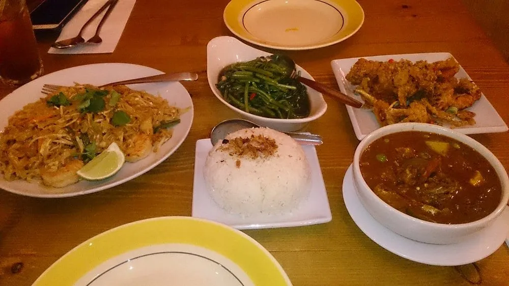 A shared table of pad thai with prawns and lime, stir-fried morning glory, fried soft-shell crab, jasmine rice and a dark curry, on a pine table with yellow-rimmed plates