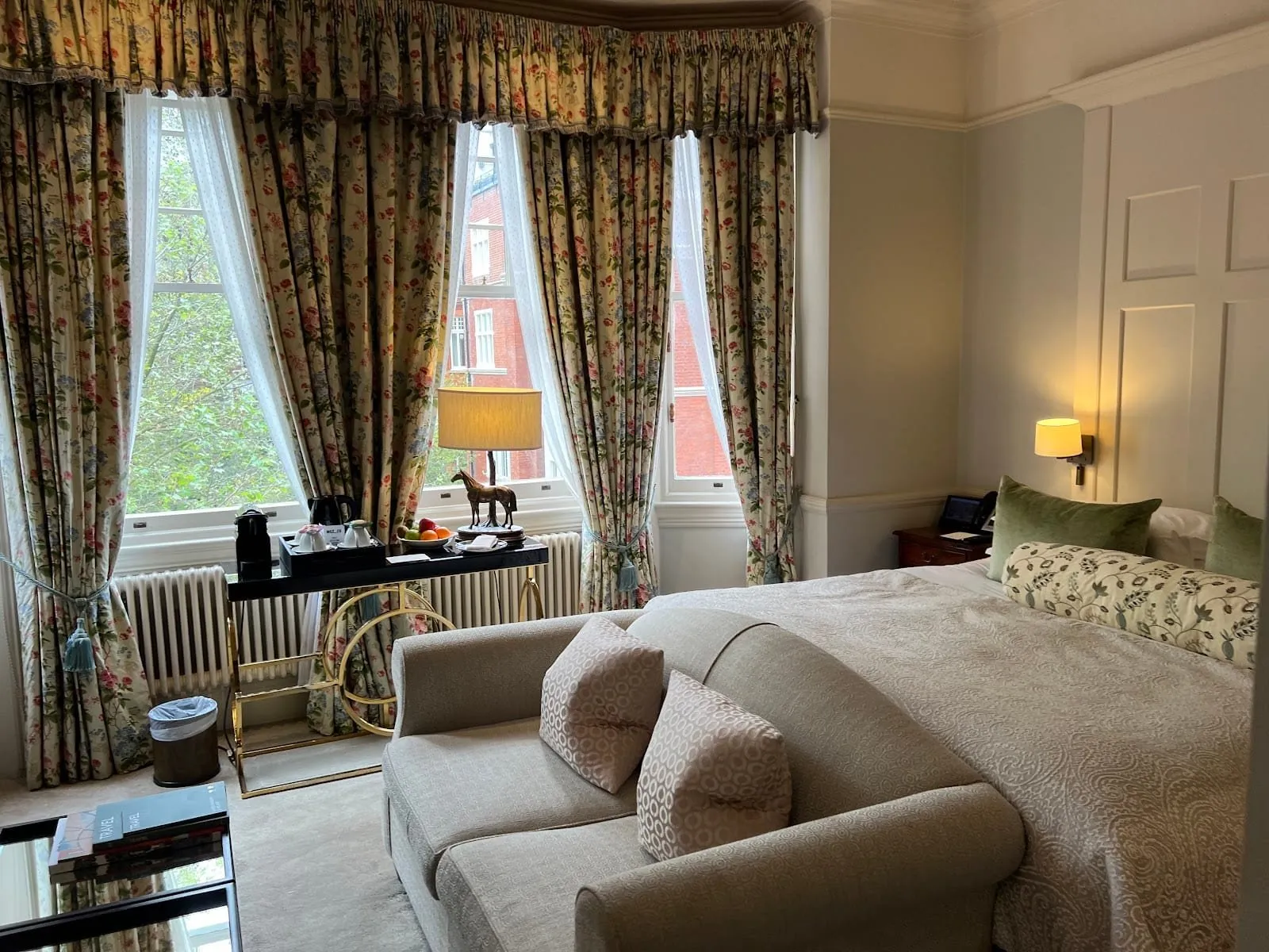 A room at 11 Cadogan Gardens with a bay of three sash windows hung with floral chintz curtains, a sofa at the foot of the bed, a coffee machine on a black and brass console, and red-brick mansion blocks outside