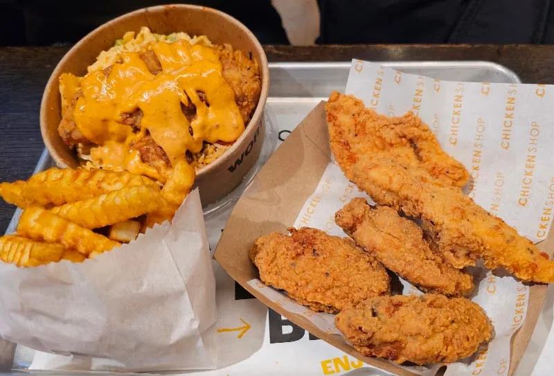 Fried chicken tenders on branded chicken shop paper beside crinkle-cut fries and a rice bowl under a thick orange sauce, on a metal tray