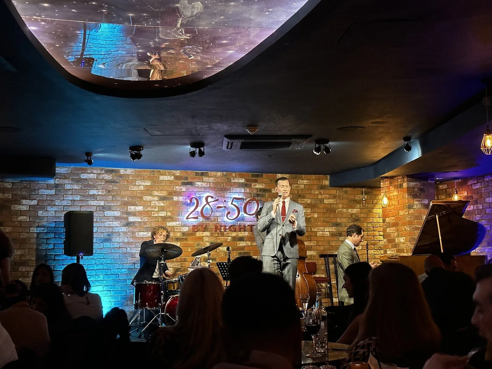 A singer performs with a drummer, double bass and piano in front of the 28-50 By Night sign, with diners in the foreground