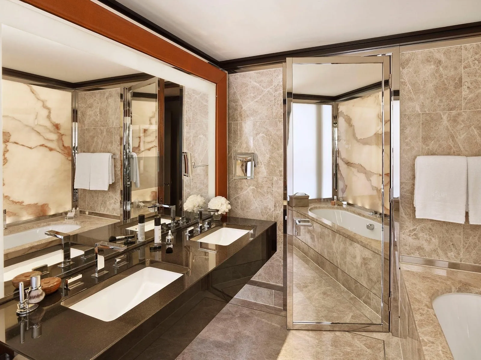 A 45 Park Lane bathroom in pale grey marble with twin basins on a dark stone counter, a bath, and an onyx panel lit from behind