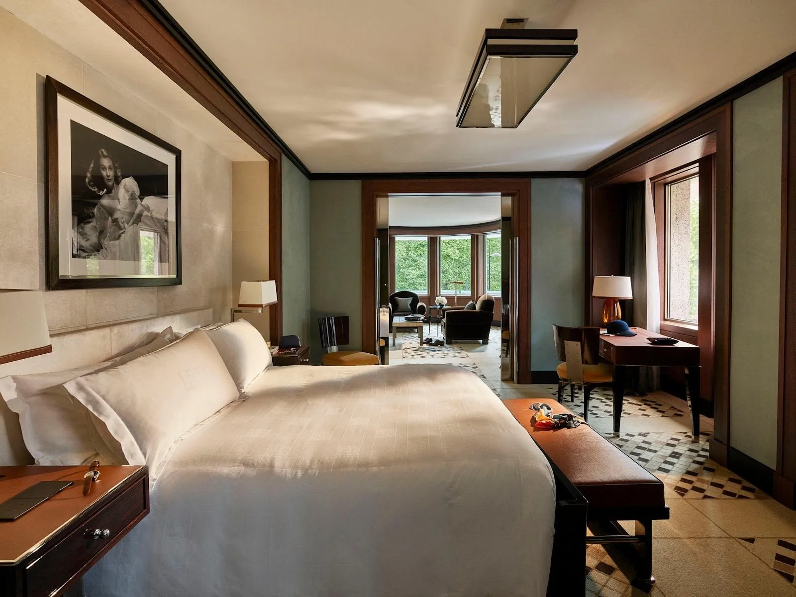 A 45 Park Lane suite: a king bed under a black-and-white film-star photograph, dark wood trim and a patterned deco carpet, with a sitting room beyond whose curved bay windows look into the trees of Hyde Park