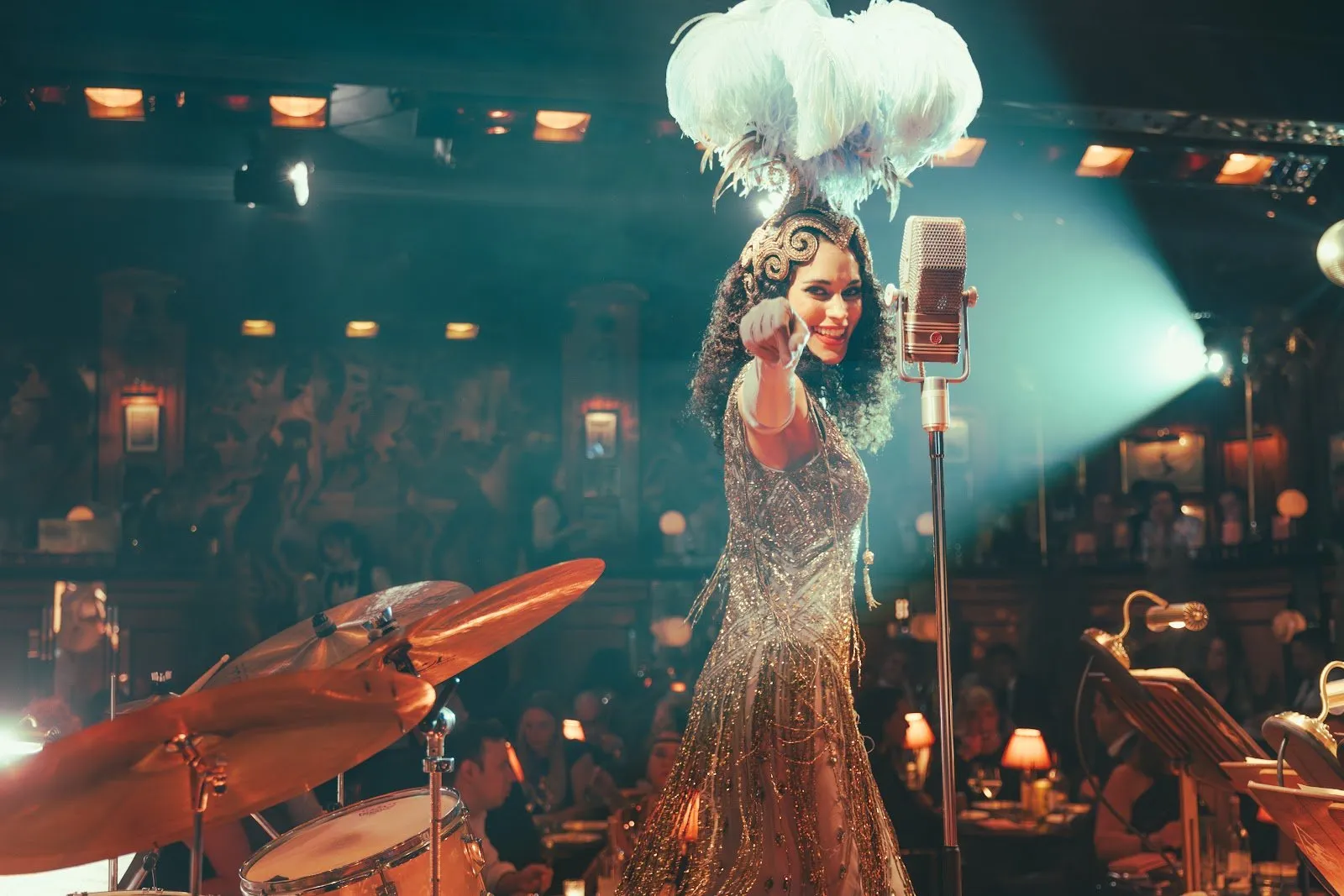 A singer in a feathered headdress and beaded 1920s dress performs at a vintage microphone, with diners at lamplit tables behind