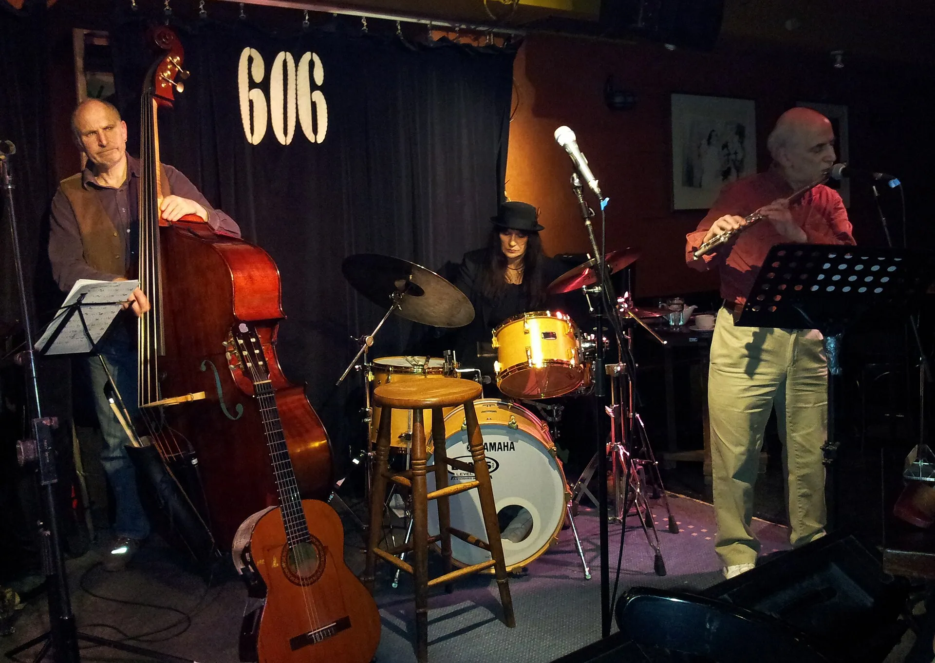 Three musicians playing in the low, close room of the 606 Club, a double bass, drums and a saxophone under stage lighting