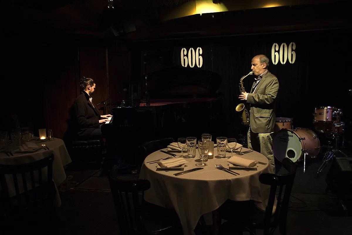 A pianist and a saxophonist play in front of the lit 606 sign, beside round tables laid with white cloths and candles