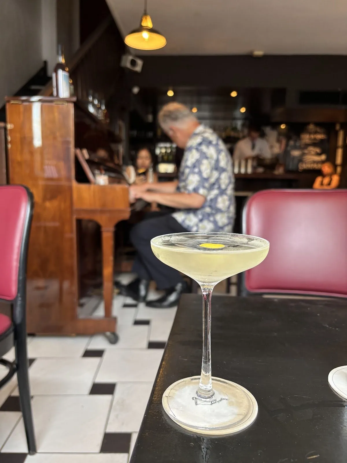 A cocktail in a coupe on a black table at 69 Colebrooke Row, with a man playing an upright piano behind