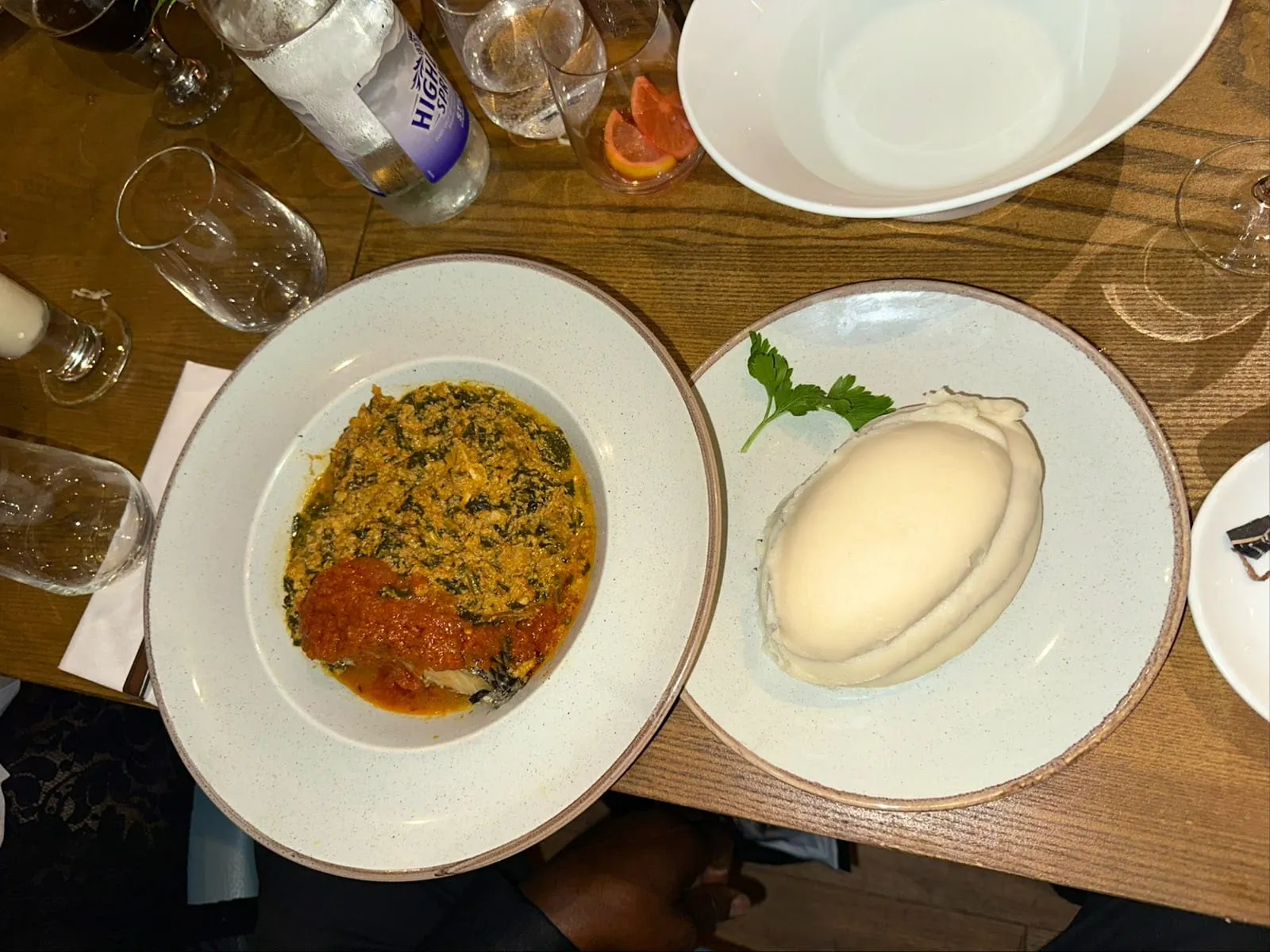 Two plates seen from above: a green leaf stew, thick and flecked, with a spoonful of red pepper sauce over it, and a smooth white oval of pounded yam on a plate of its own