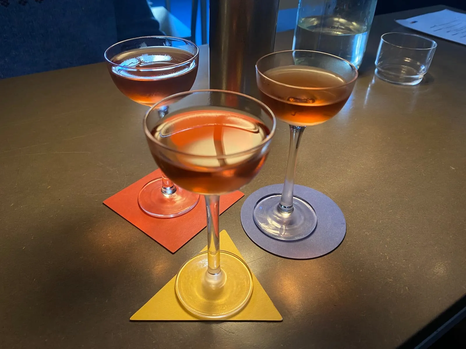 Three amber cocktails at A Bar with Shapes for a Name, on coasters cut as a yellow triangle, a red square and a blue circle