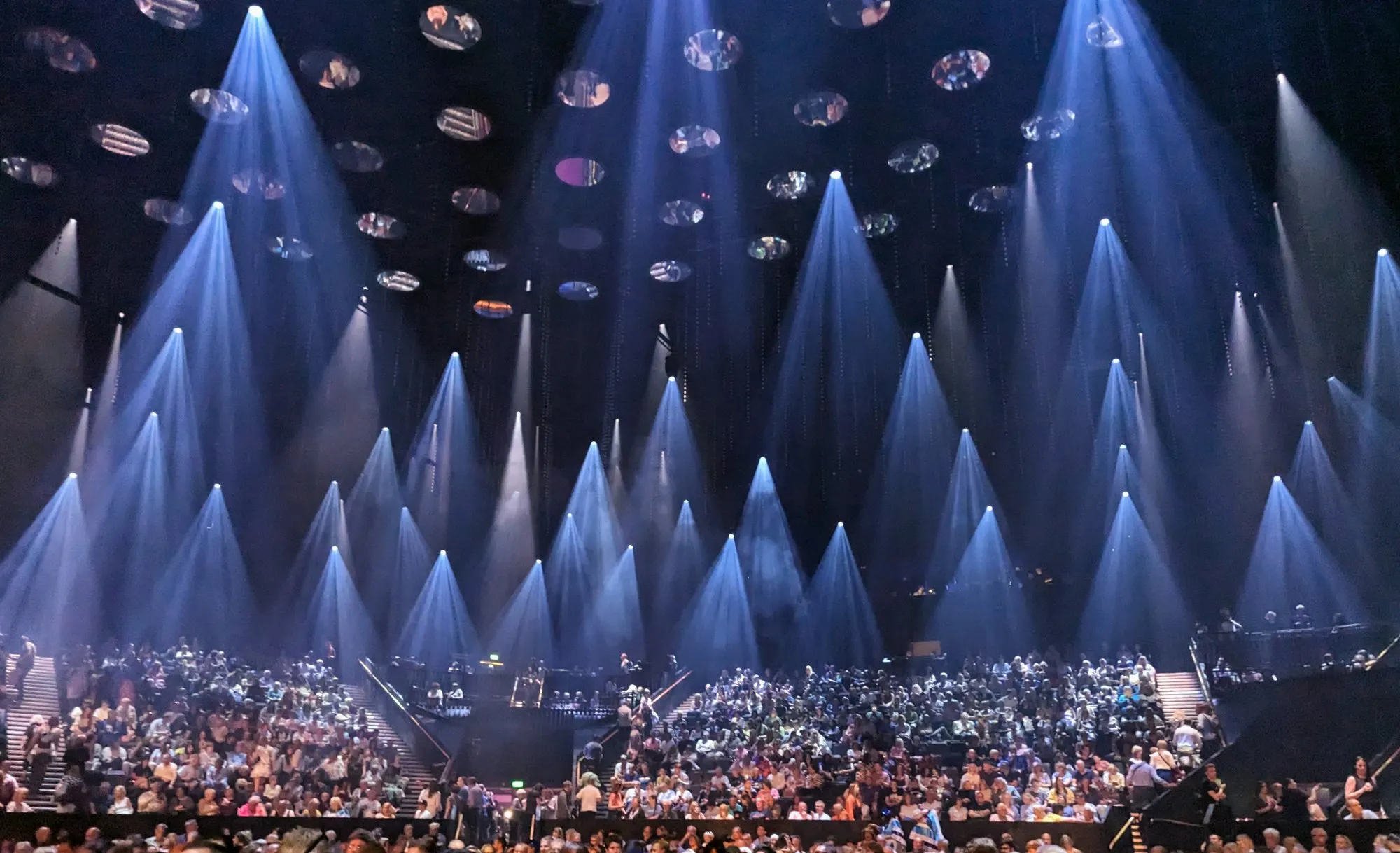 Dozens of narrow blue light beams angled down over a full audience inside the ABBA Arena before the show