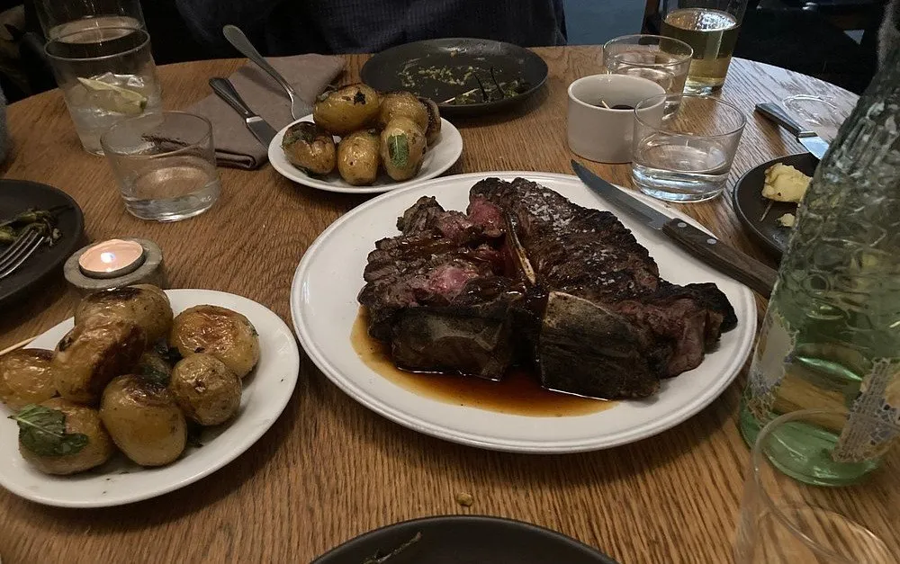A sliced T-bone steak resting in its juices on a white plate, with two dishes of roast new potatoes and herbs on a wooden table