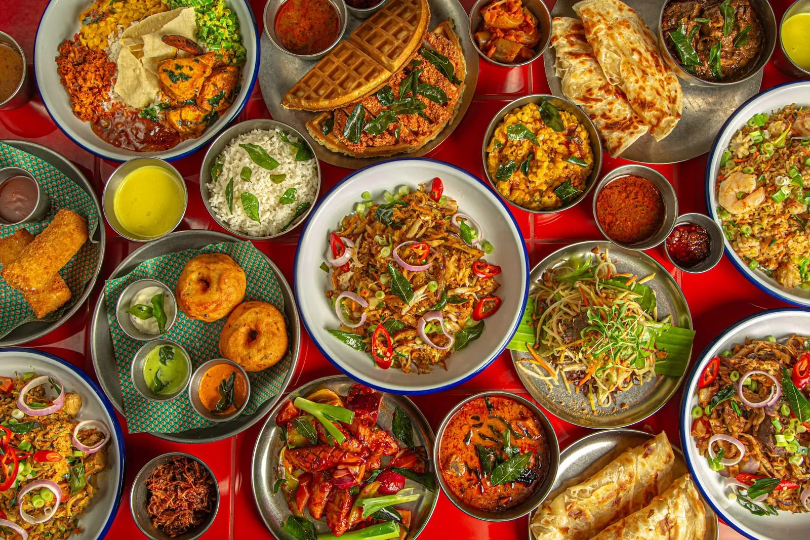A bright red table crammed with dishes: fried chicken laid over a golden waffle with fried curry leaves, blue-rimmed bowls of kottu topped with red onion rings and chilli, flaky folded roti, two lentil vadai with three chutney pots, dal studded with curry leaves, a bowl of rice scattered with prawns, and a steel pot of dark curry under fried curry leaves
