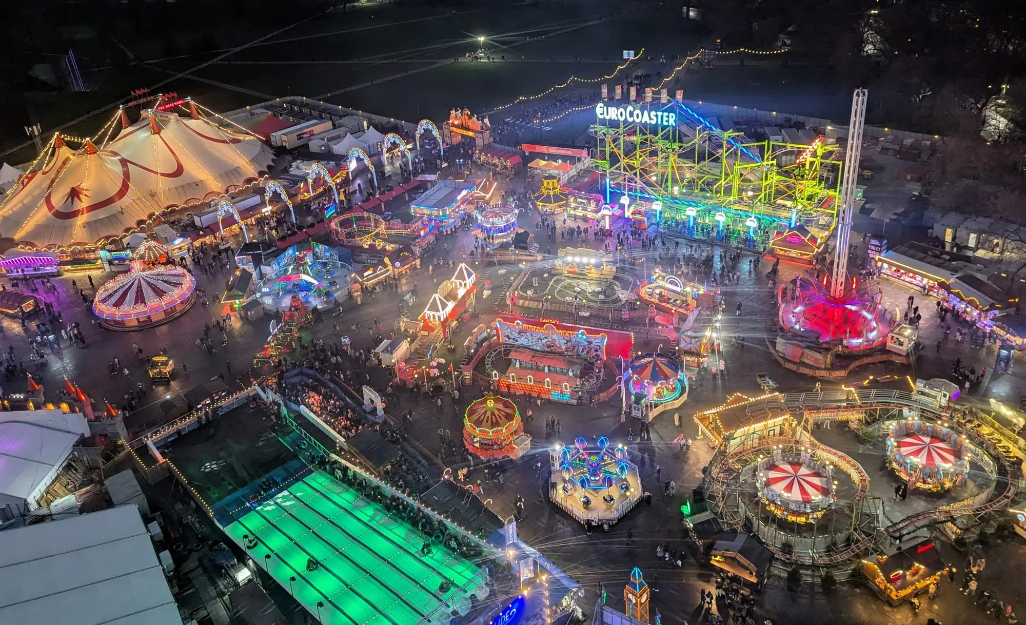 An aerial night view over Hyde Park Winter Wonderland showing the illuminated fairground, the green ice rink, the big top and the Euro Coaster