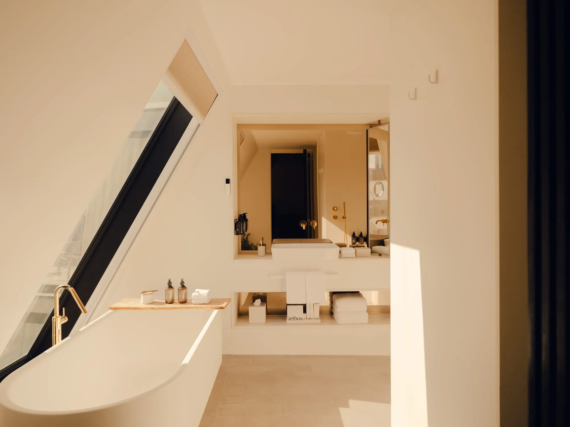 A bathroom at Aethos London Shoreditch with a free-standing tub under a sloped skylight window and gold fixtures