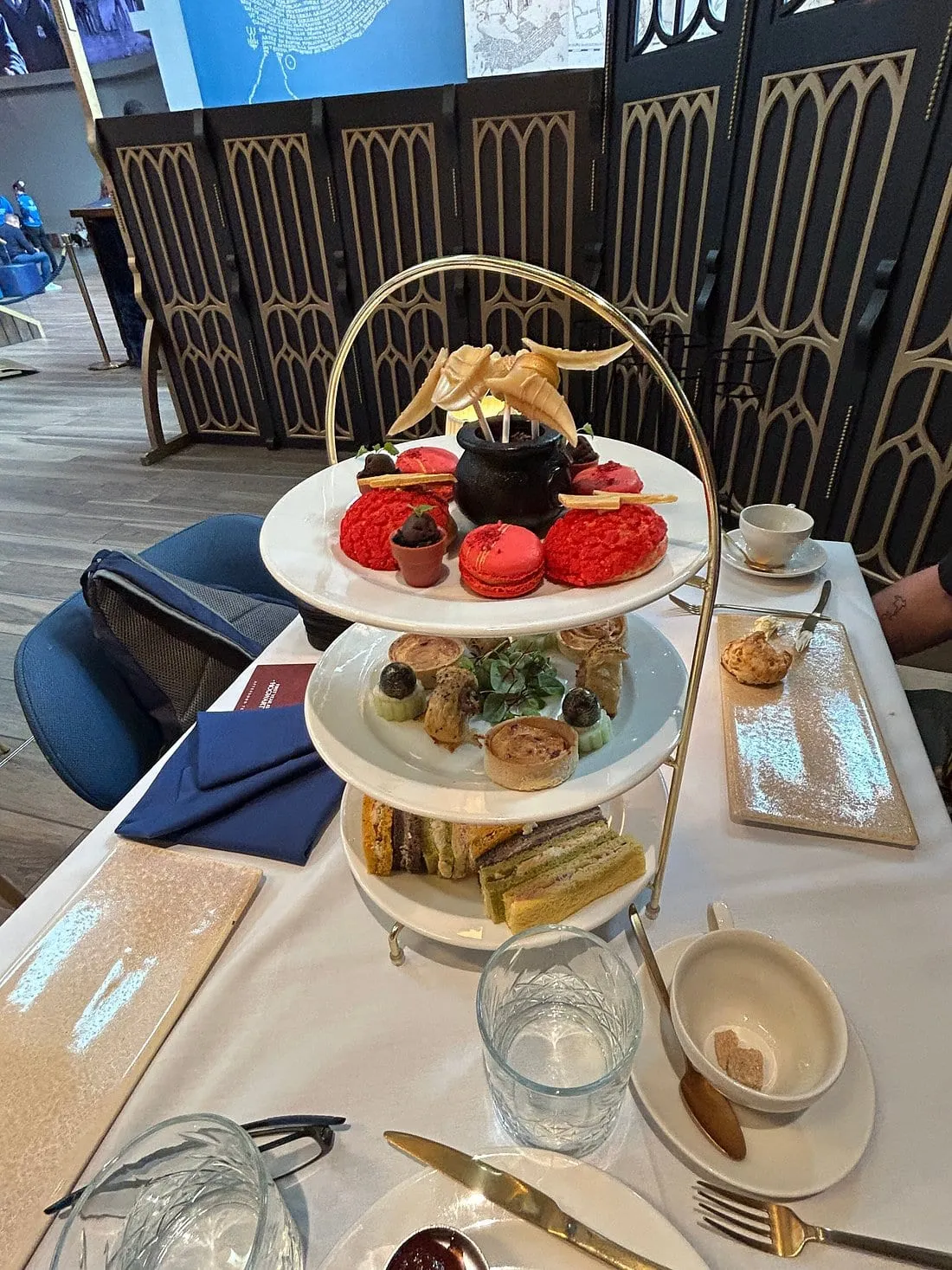A three-tier afternoon tea stand at the Studio Tour: sandwiches below, savoury tarts in the middle and red iced cakes, macarons and a chocolate cauldron on top, with a golden snitch on the handle