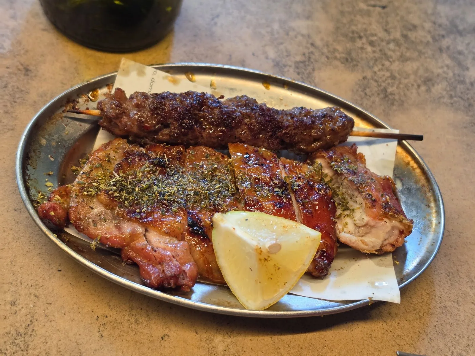 A minced lamb skewer and grilled chicken thigh scattered with oregano, with a lemon wedge, on a metal plate