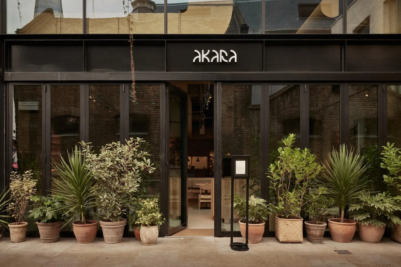 The black shopfront of Akara in Borough Yards, its name lit above folding glass doors, with terracotta pots of shrubs and palms lined along the pavement and a Stoney Street sign at the left edge