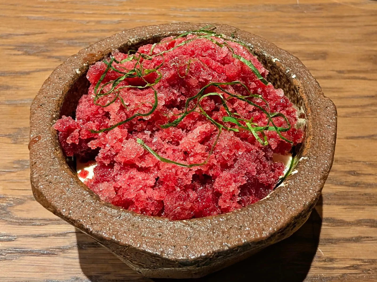 A coarse red granita heaped over a pale cream base in a rough hand-thrown stone bowl, scattered with fine green shreds of leaf