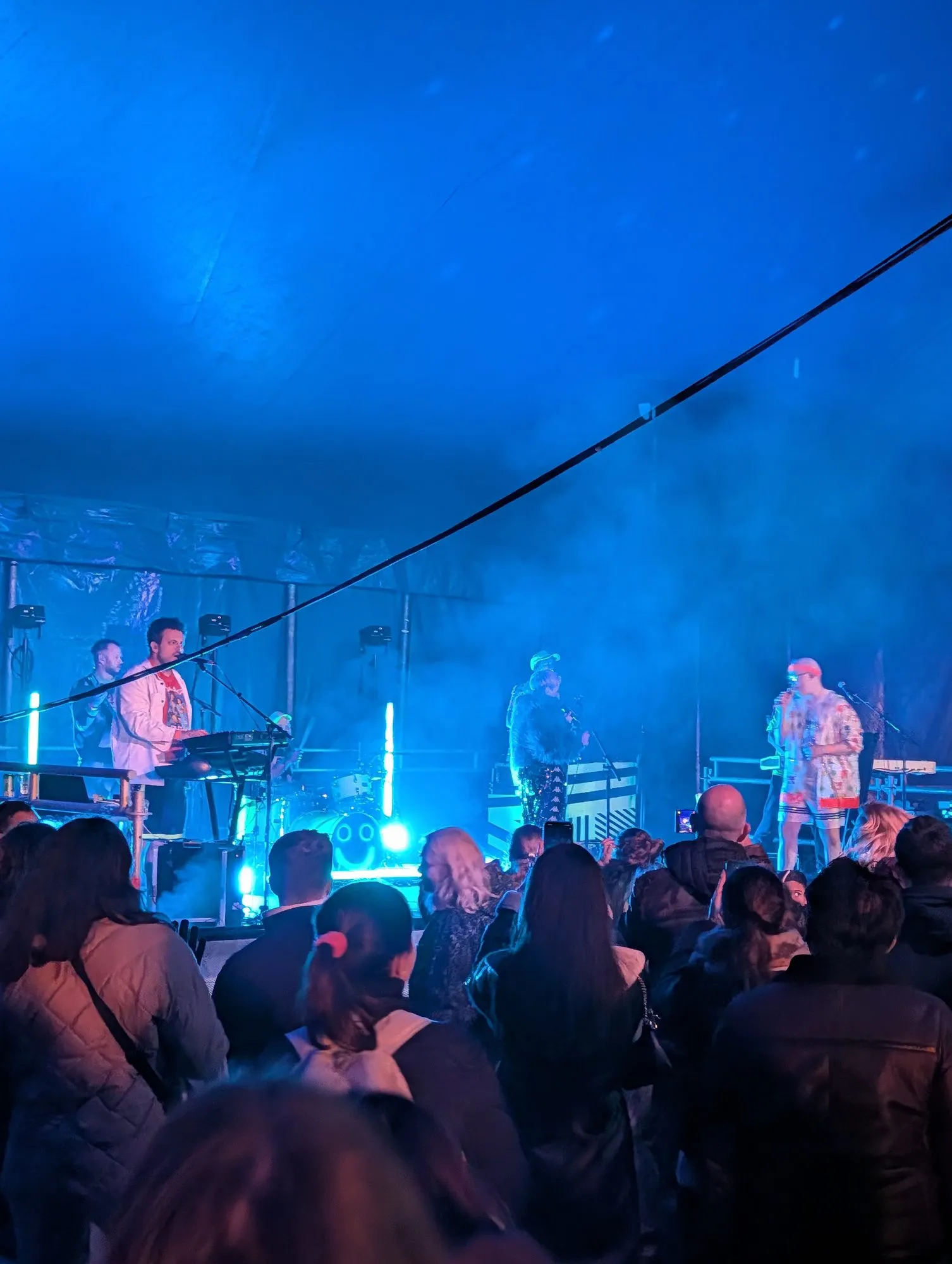 A band playing on a stage inside a marquee lit blue, with a crowd watching