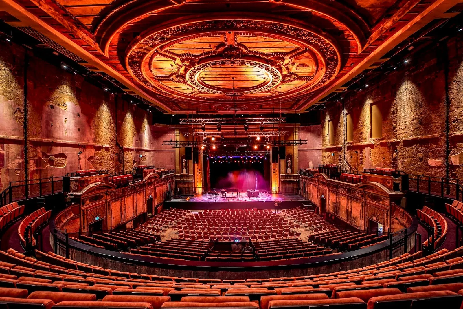 The theatre at Alexandra Palace, with rows of orange seats in the stalls and circle, bare peeling walls and an ornate plaster ceiling lit in orange