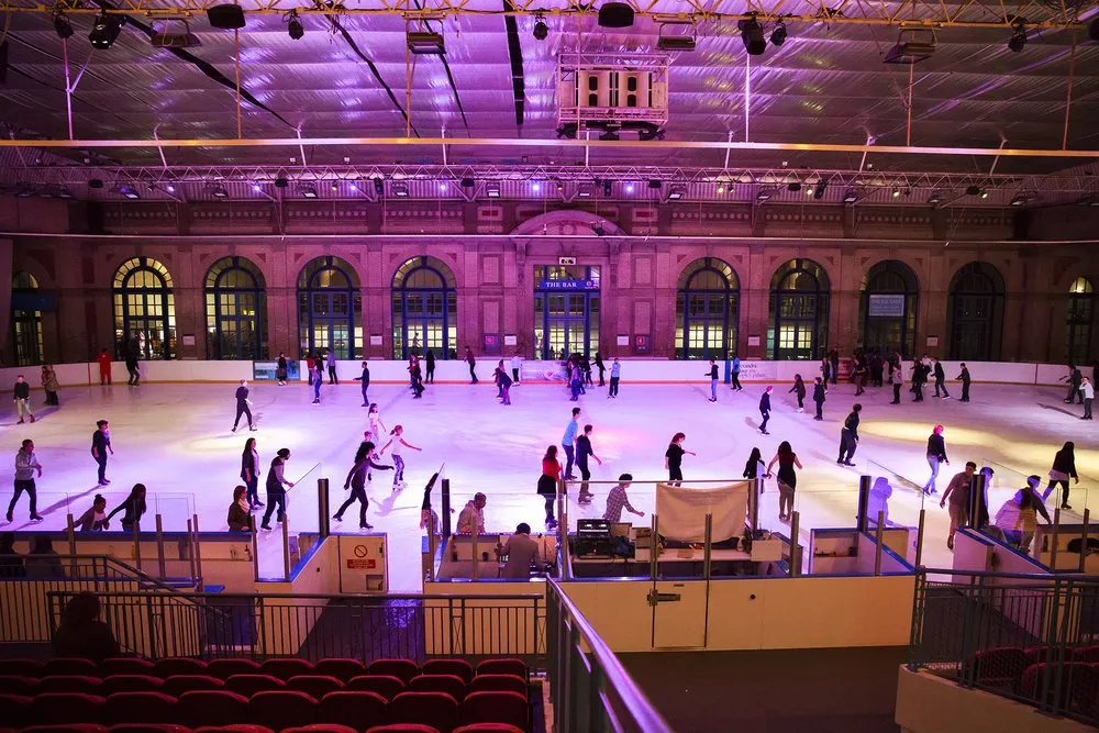 Skaters on the indoor Alexandra Palace ice rink under pink and purple lights, the hall's arched Victorian windows along the far side