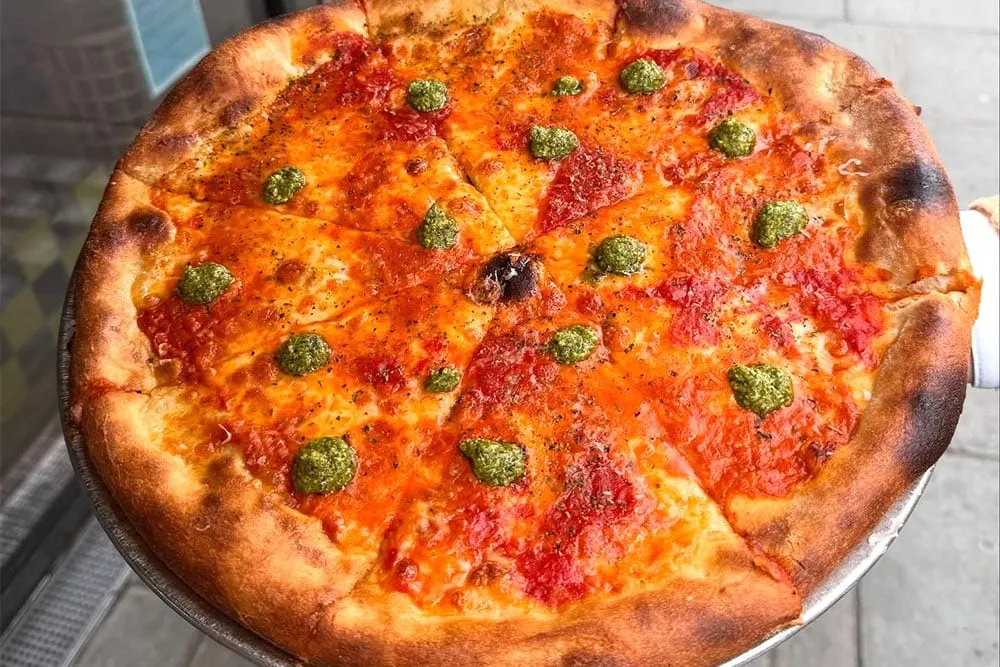 An Alley Cats pizza cut into slices on a metal tray, tomato sauce and thin melted cheese under dollops of green pesto and a scatter of dried oregano, held up on the pavement outside