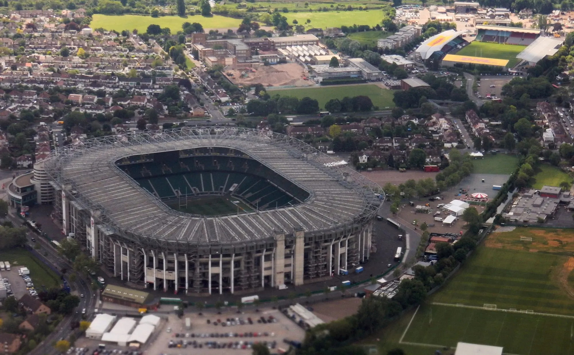 Twickenham's stadium bowl from the air, its steel roof trusses open over the pitch, with the Twickenham Stoop and the A316 beyond it and terraced streets on every other side