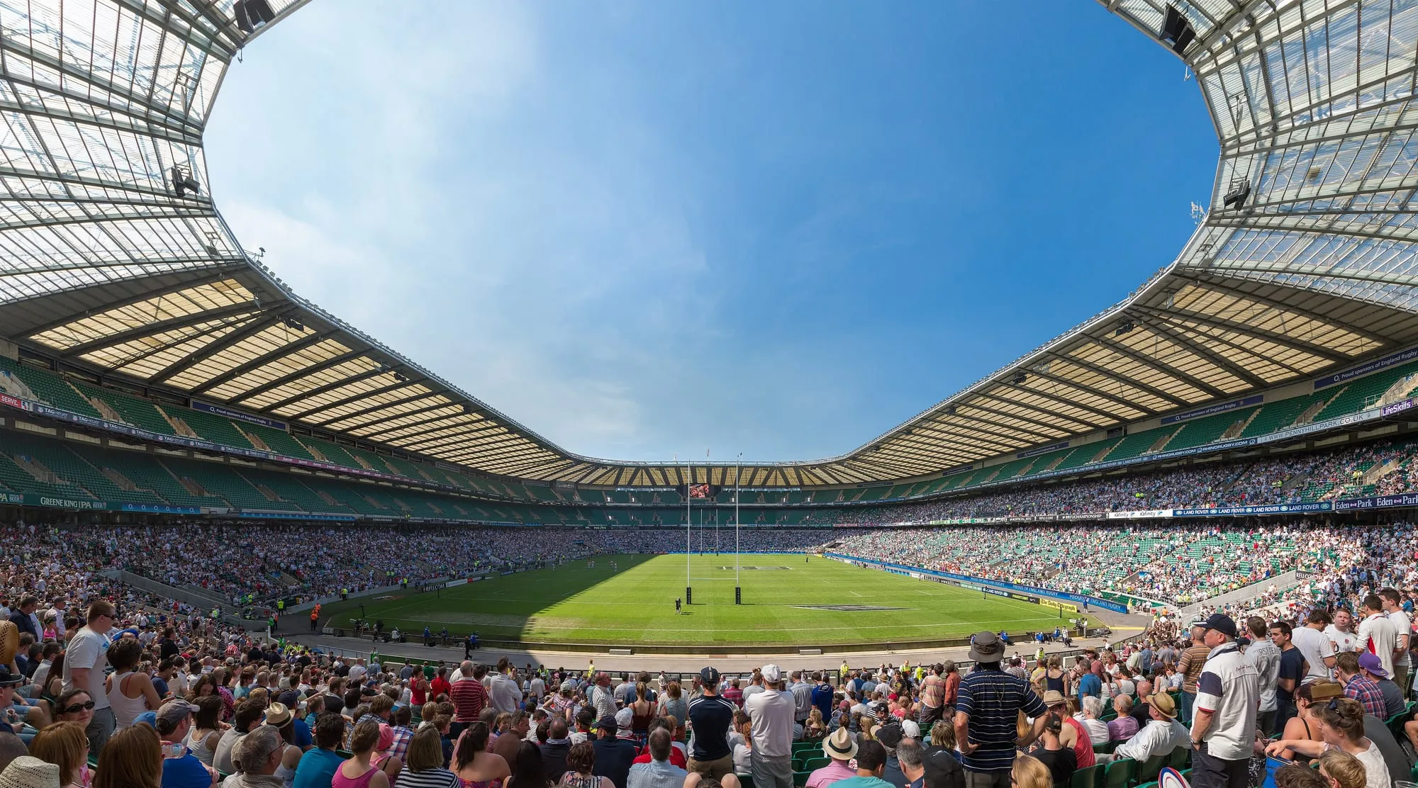 Twickenham's seating bowl from high in a corner: a sunlit pitch below three tiers of green seats, the crowd in, and blue sky through the open roof