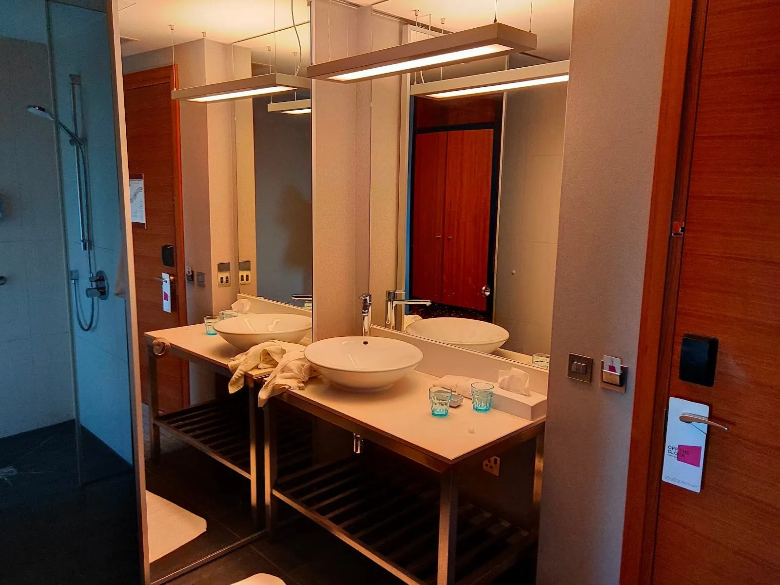 A vessel basin at Aloft London ExCeL on a white counter with an open slatted shelf below, a long mirror behind and a side mirror doubling it, by the room door and a glass-screened shower