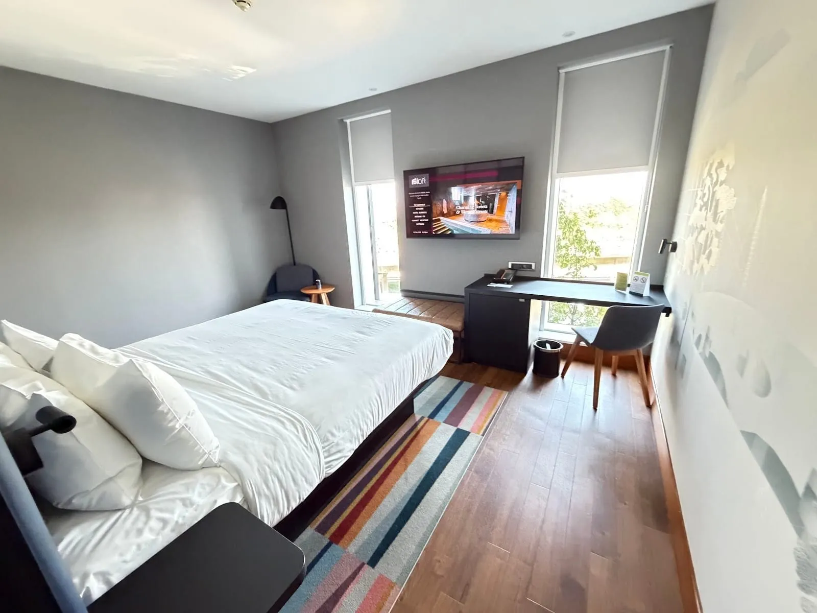 A room at Aloft London ExCeL with a double bed, grey walls, a striped runner rug on a wood floor, a wall-mounted TV between two tall windows with blinds, and a desk and chair under the right-hand window