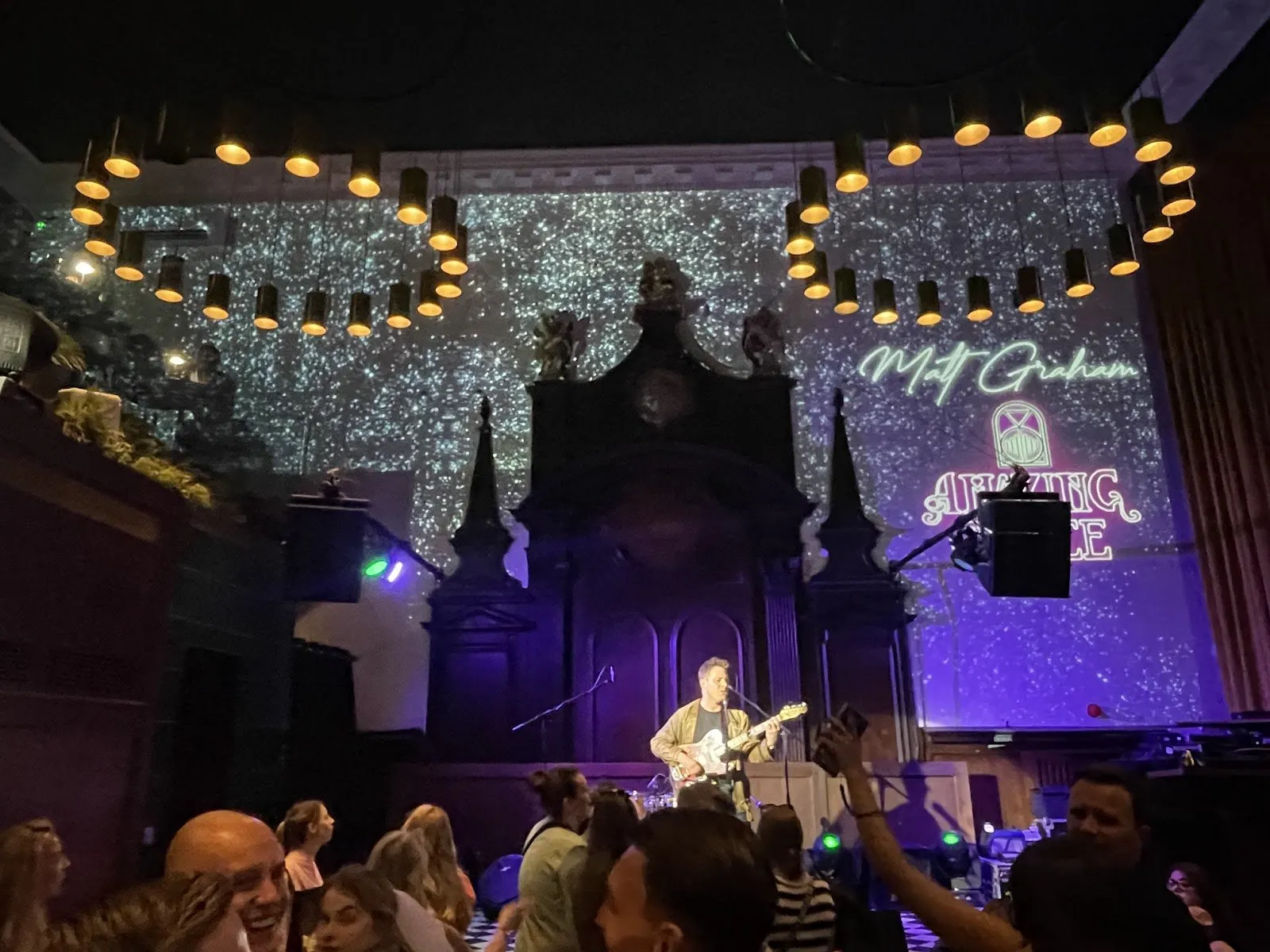 A guitarist sings on the stage of Amazing Grace, a former church, with the venue's name projected behind him