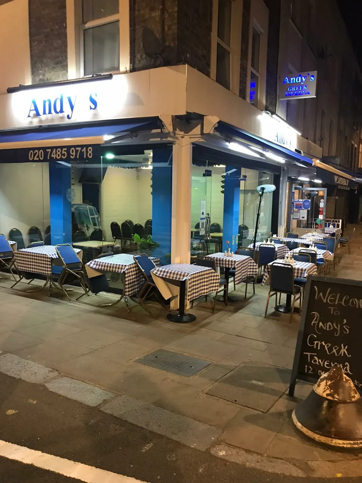 Andy's Taverna at night, a corner restaurant with a blue-and-white fascia, checked tablecloths on pavement tables and a chalkboard reading Welcome to Andy's Greek Taverna