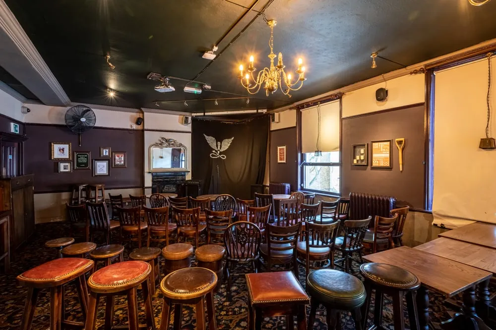 The upstairs room at The Camden Head set for comedy, rows of wooden pub chairs and leather stools facing a black curtain with an angel emblem