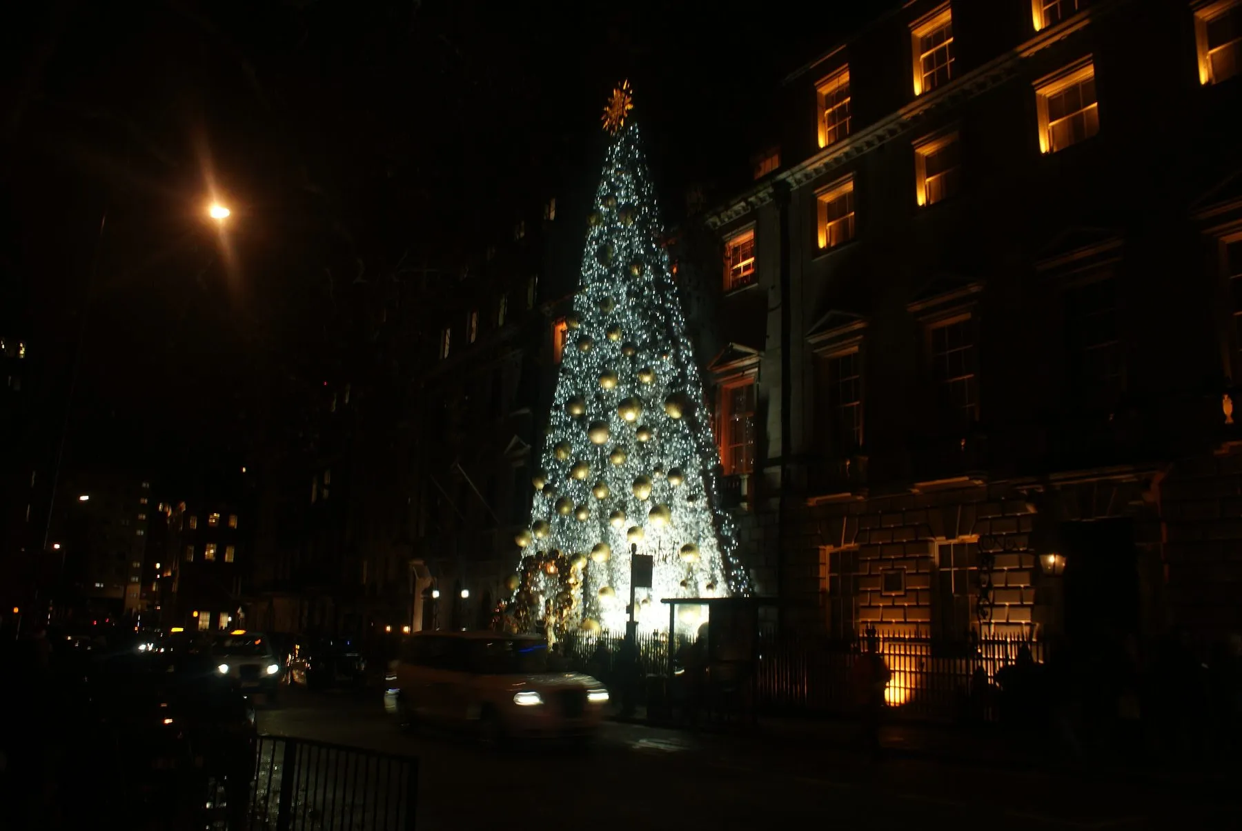 A lit Christmas tree hung with gold baubles covering the front of Annabel's at night on Berkeley Square, with a black cab passing in the foreground