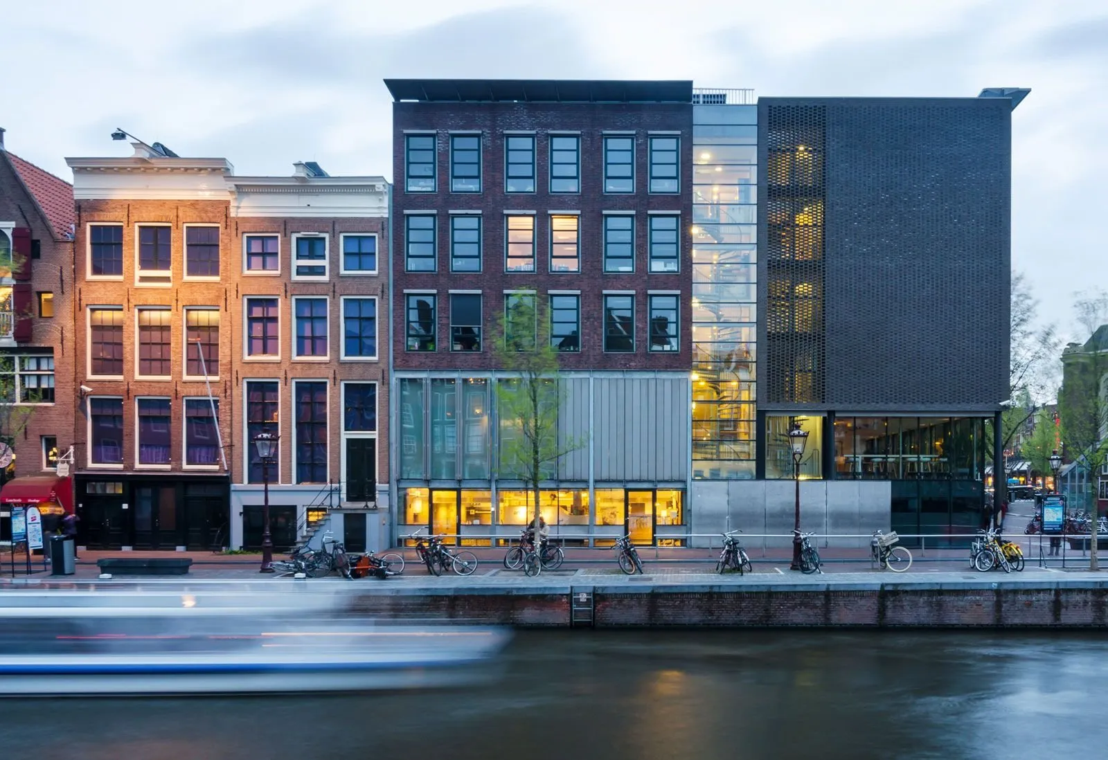 The Anne Frank House's modern black entrance building beside the canal at dusk, with the original gabled canal house it adjoins lit up next door