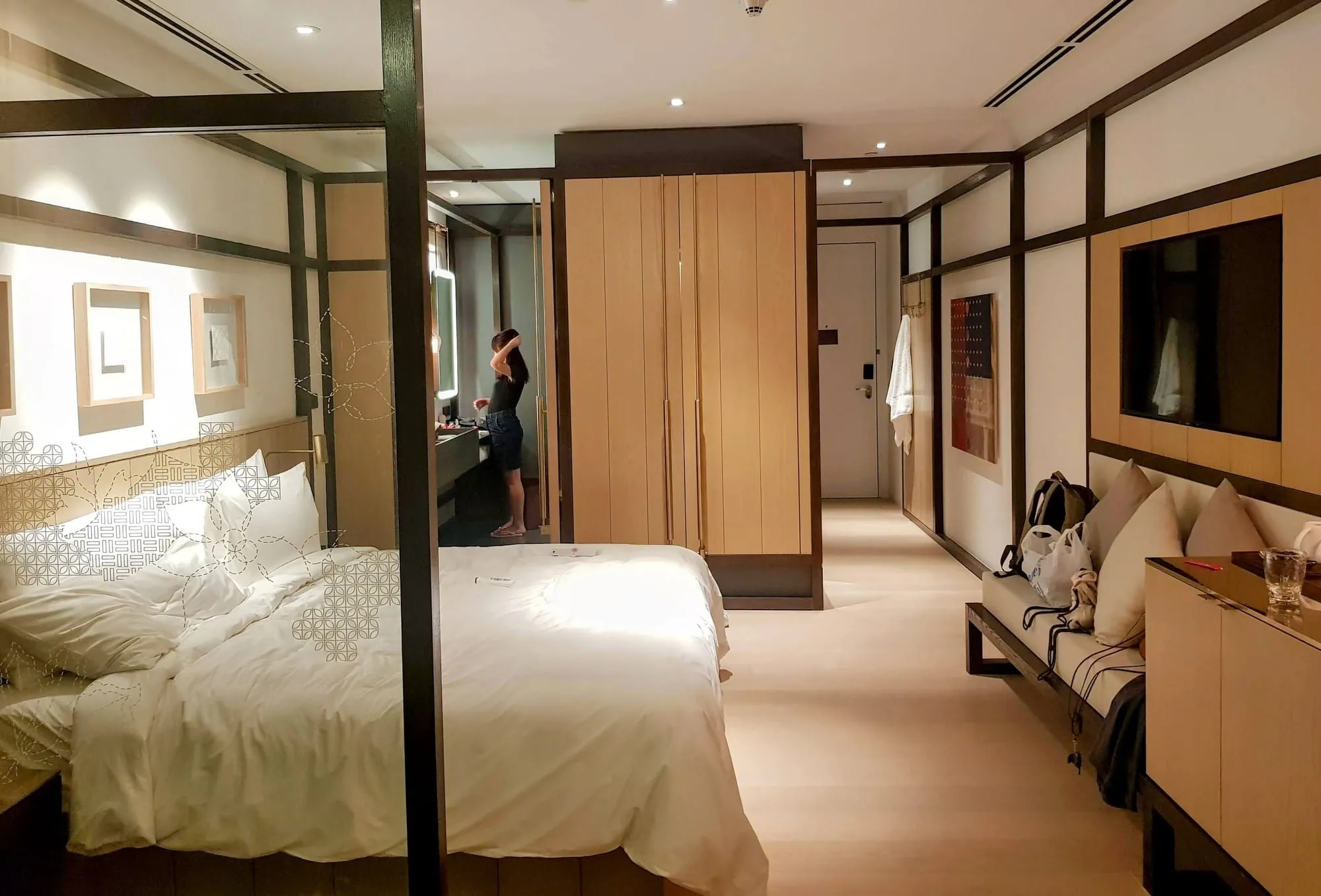 A studio room with a bed, a sitting area with a bench sofa and cushions, a full-height wardrobe and a screened bathroom beyond