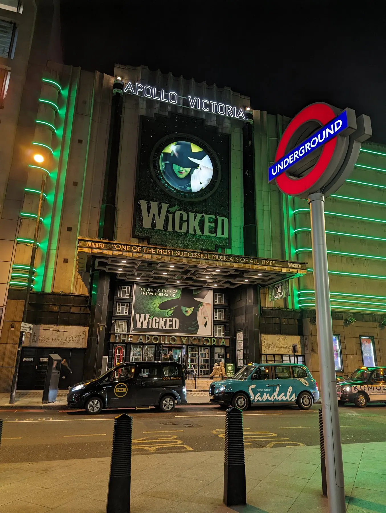 The Art Deco frontage of the Apollo Victoria Theatre lit green at night for Wicked, with black cabs and an Underground sign in front