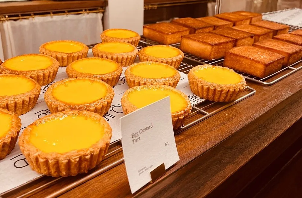 Rows of egg custard tarts with glossy yellow filling in fluted pastry cases, on wire racks on a wooden bakery counter
