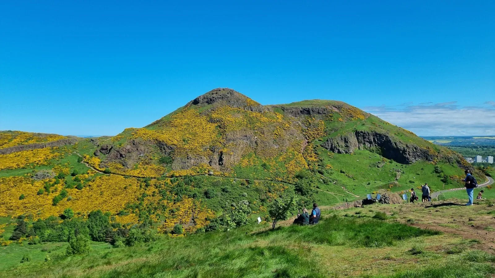 Arthur's Seat rising above gorse-covered slopes, with walkers on the path up and the city visible in the distance