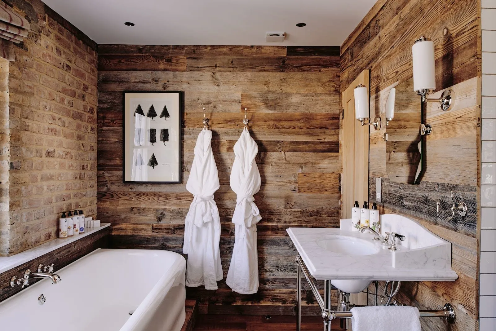 A bathroom at Artist Residence London lined with reclaimed timber and bare brick, with a bath, a marble washstand on chrome legs and two white robes on hooks