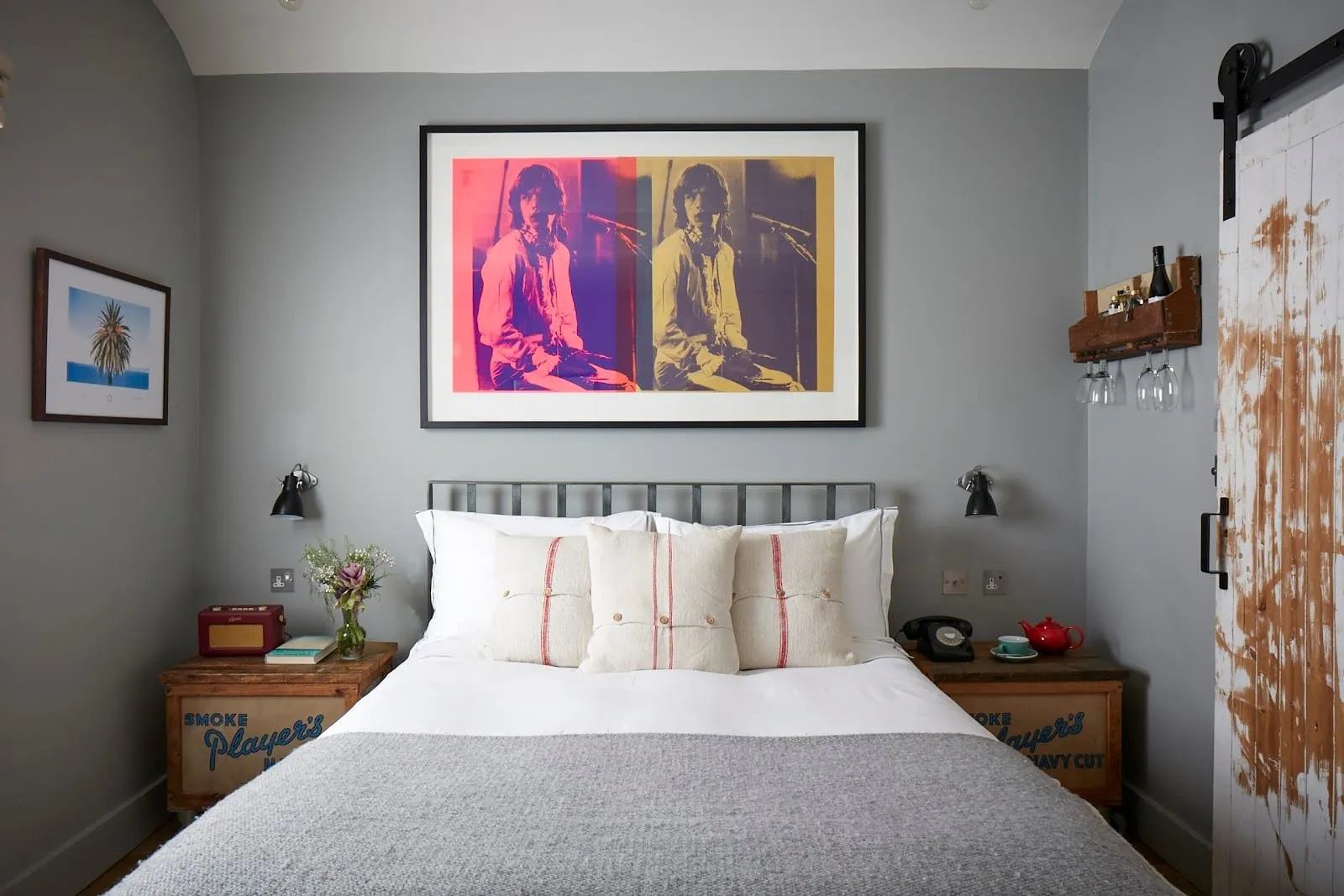 A double room at Artist Residence London with grey walls, an iron bedstead, old Player's cigarette crates as bedside tables, a pop-art print above the bed and a sliding barn door of weathered white planks