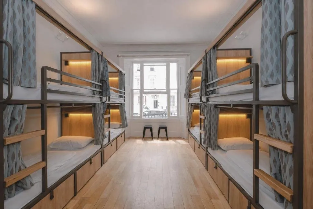 A dorm at Astor Victoria with two rows of metal-framed bunks, each with a privacy curtain, a reading light and storage drawers underneath, on a wooden floor leading to a sash window