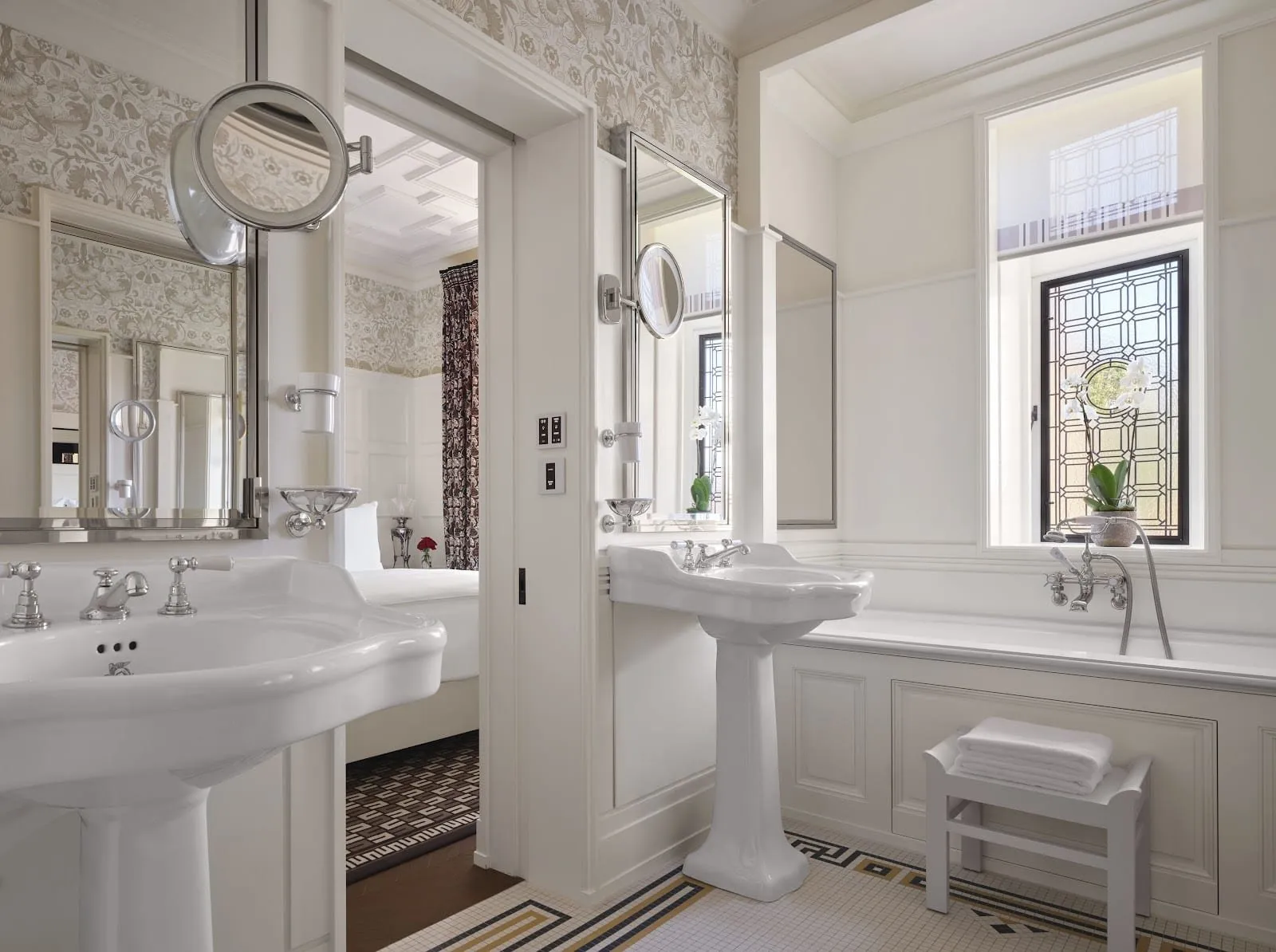 A bathroom at the At Sloane hotel with two pedestal basins, a panelled bath with a hand shower under a leaded-glass window, floral wallpaper and a mosaic floor with a Greek key border