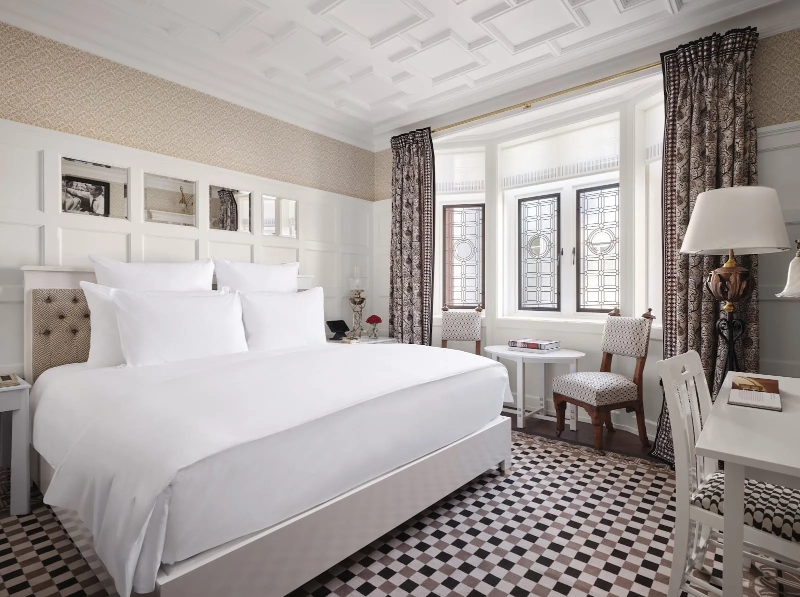 A bedroom at the At Sloane hotel with white panelled walls and mirrored panels above the bed, a coffered ceiling, a chequered black, brown and white carpet, patterned curtains and a bay of leaded-glass windows