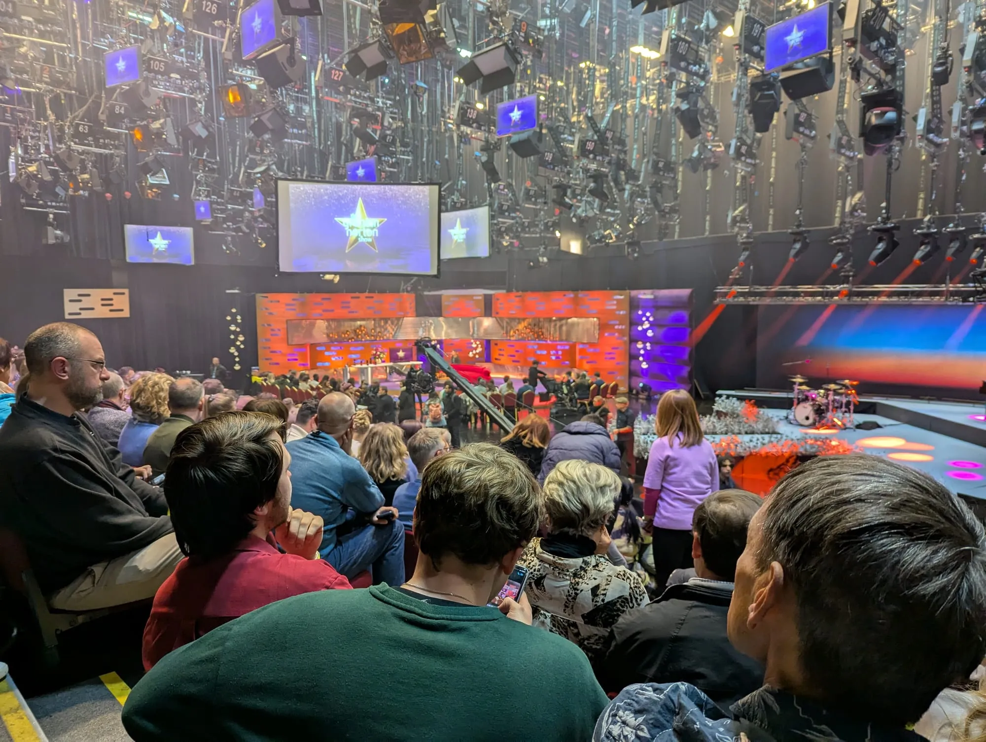 The seated audience waiting in the studio before recording begins, with crew moving equipment around the set and a camera crane in position