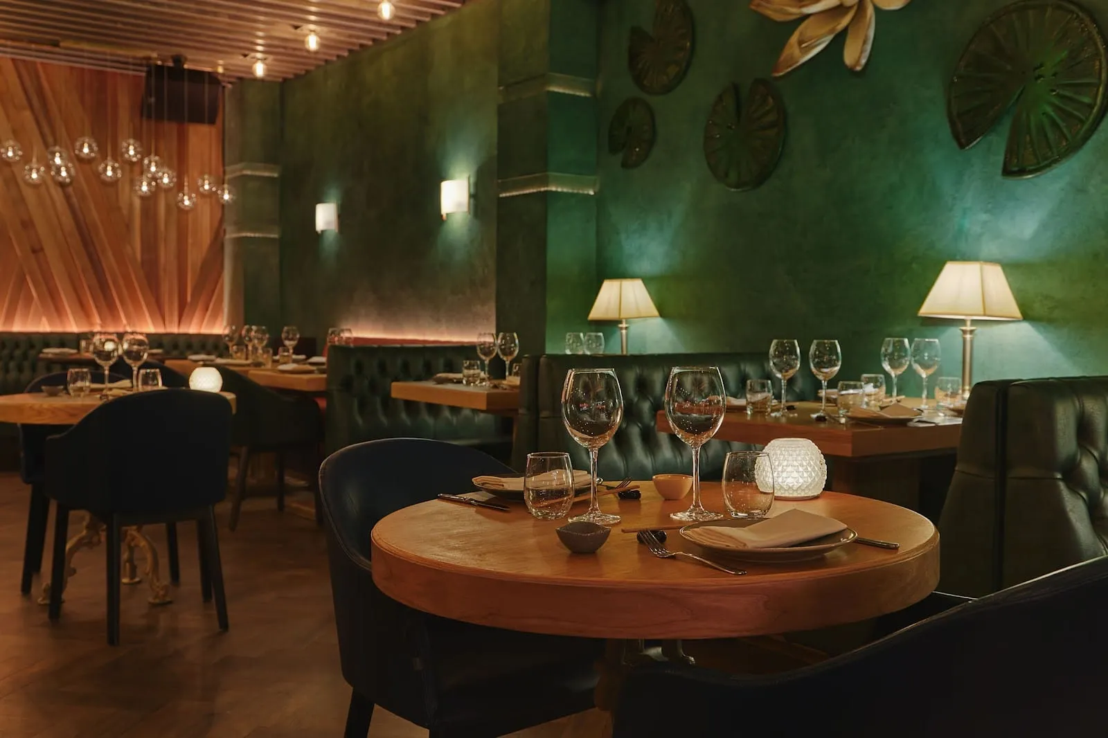 A dark green dining room with lily-pad reliefs on the wall, buttoned green leather banquettes, navy tub chairs, round wooden tables and cream table lamps