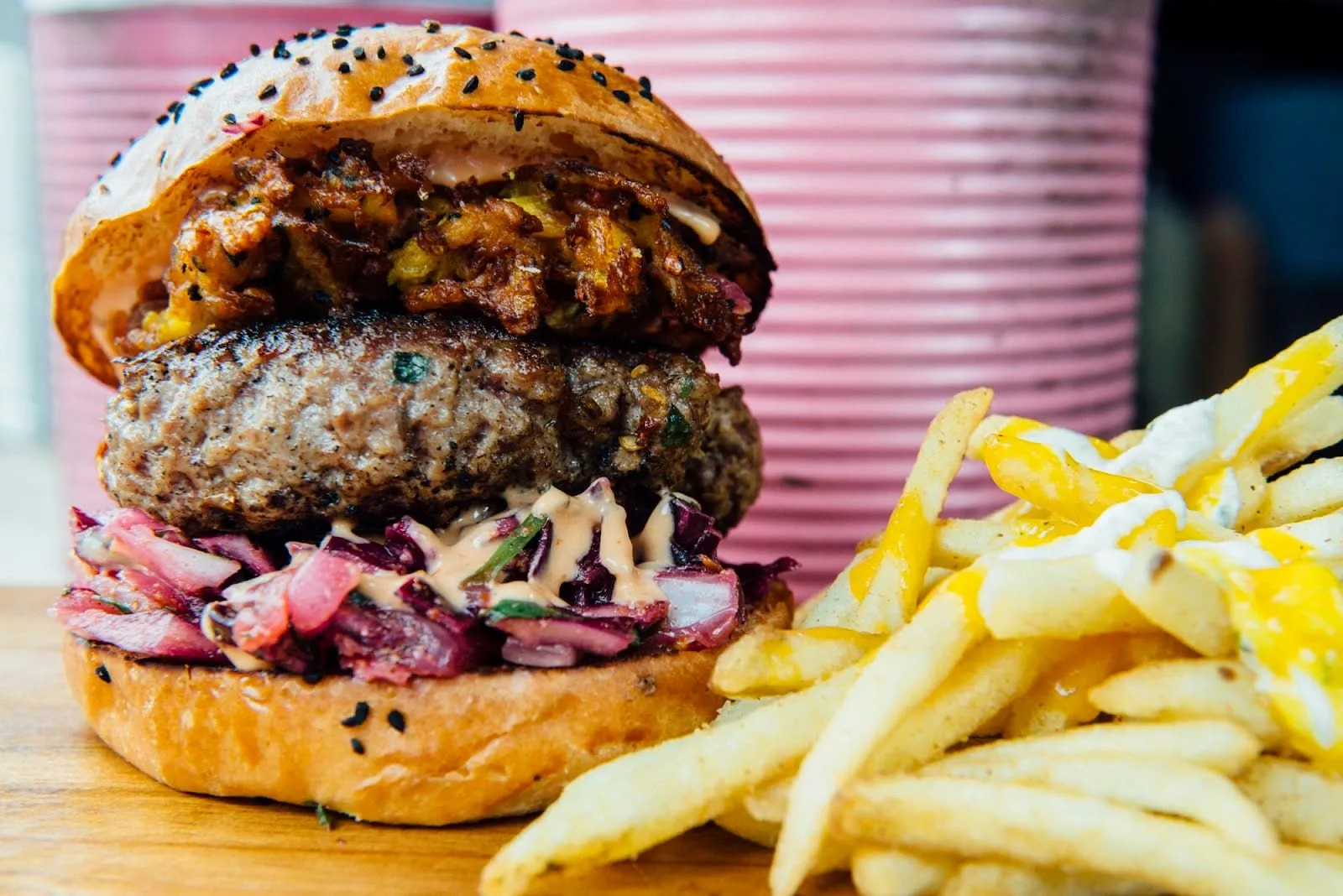 A Baba G's burger beside a pile of fries: a thick, herb-flecked patty on pink pickled slaw and sauce, topped with a crisp fried fritter, in a bun scattered with black seeds