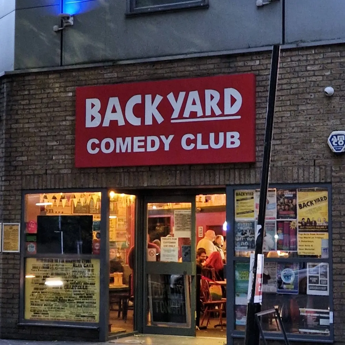 The red Backyard Comedy Club sign above a lit-up brick shopfront in the evening, with posters in the windows