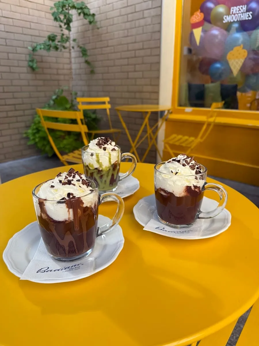 Three glass mugs of thick Badiani hot chocolate topped with whipped cream and chocolate shavings, on branded napkins at an outdoor table