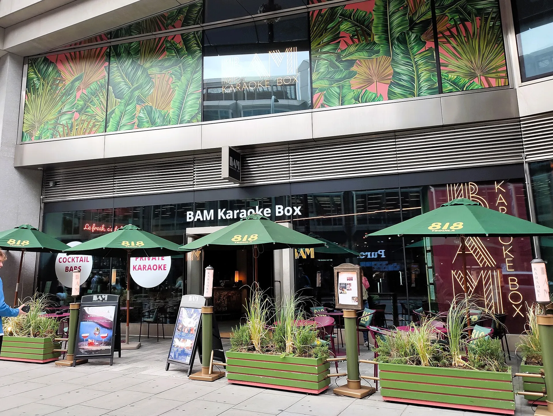 The glass front of BAM Karaoke Box on Victoria Street, with green parasols over a pavement terrace, signs reading Private Karaoke and Cocktail Bars, and a jungle-print mural on the floor above